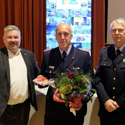Bild vergr&ouml;&szlig;ern: Das Bild zeigt 3 M�nner. Ein Mann h�lt einen Blumenstrau� und ein Geschenk in den H�nden.