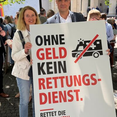 Bild vergr&ouml;&szlig;ern: Das Bild zeigt 2 Menschen, die ein Plakat halten auf dem steht "Ohne Geld kein Rettungsdienst."