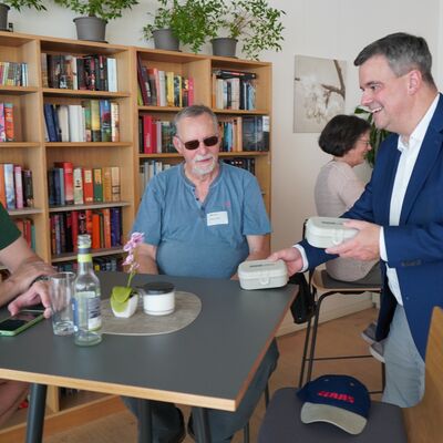 Bild vergr&ouml;&szlig;ern: Das Bild zeigt zwei M�nner, die an einem Tisch sitzen. Eine andere Person �berreicht ihnen ein Geschenk.